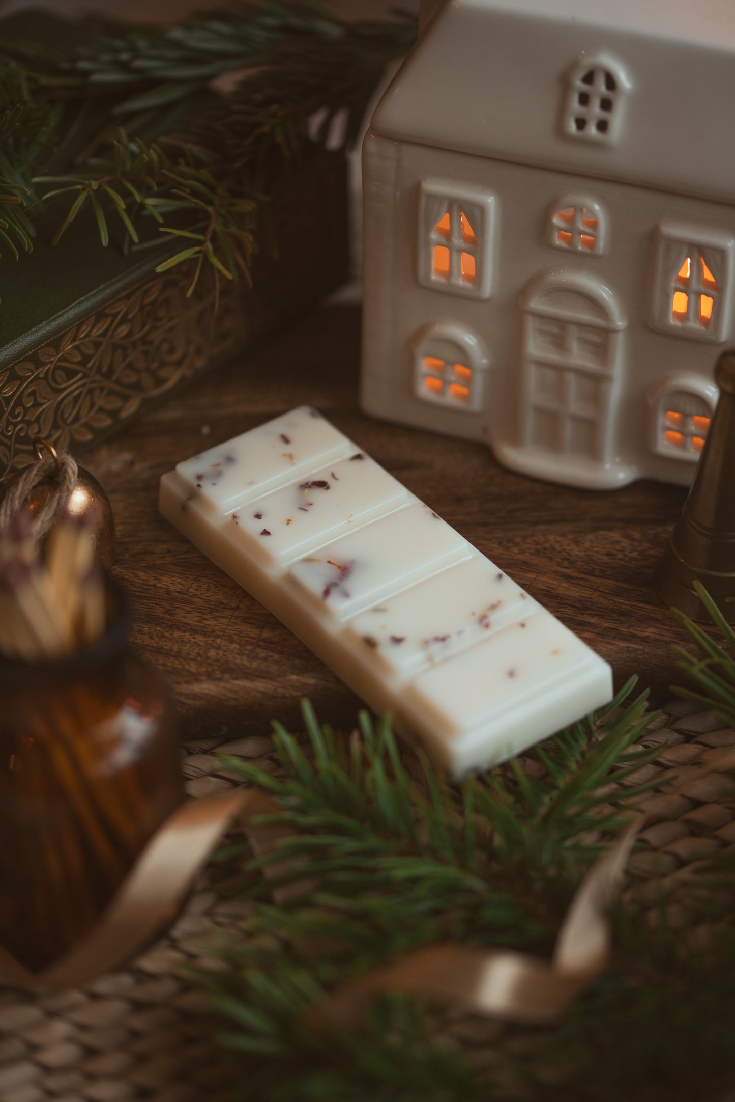 hand poured soy wax melt snap bars surrounded with twigs of fir tree and a ceramic house wax melt burner which is lit with a candle 